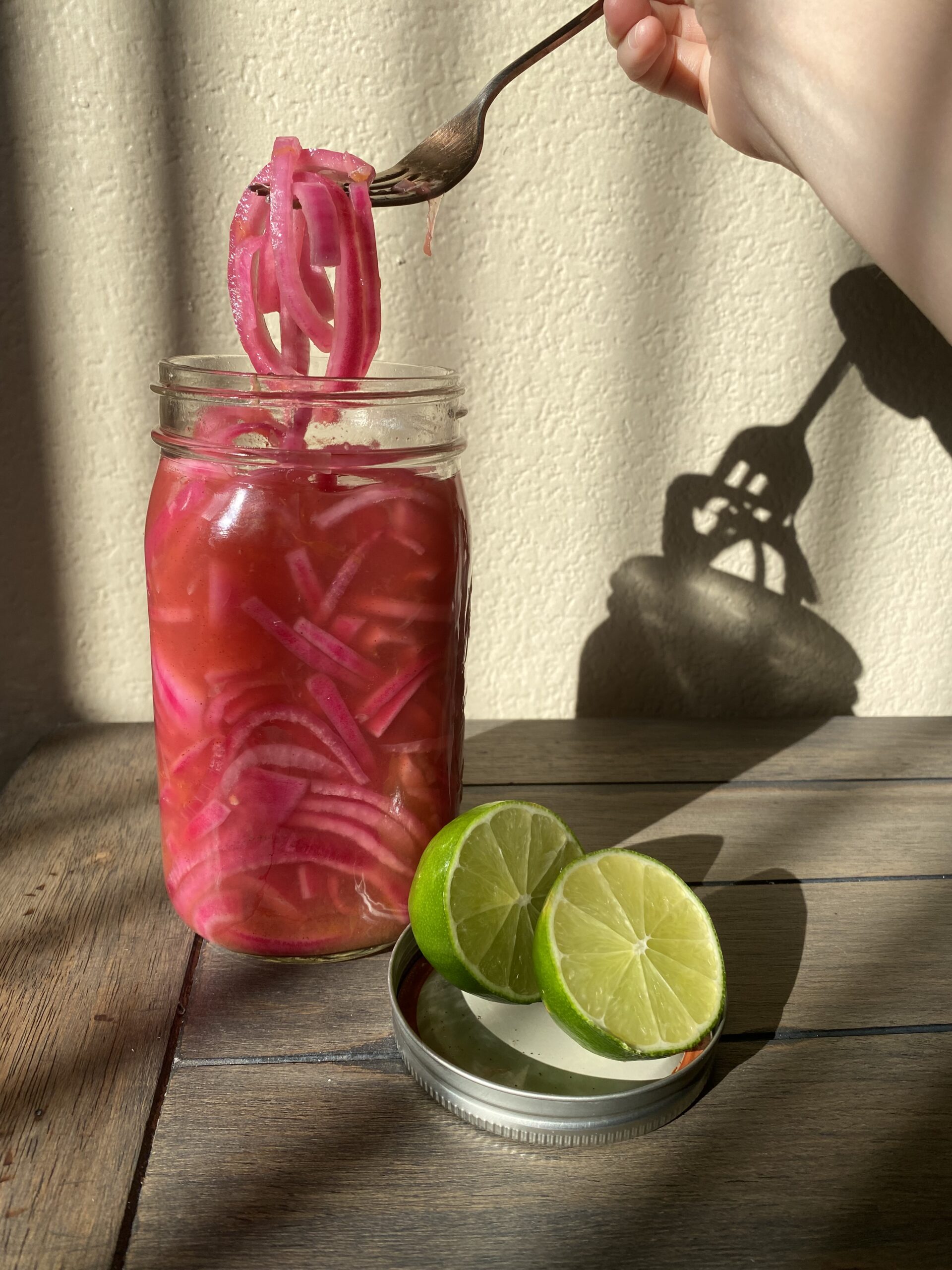 Pickled red onion being scooped out of a mason jar with a fork so you can see the onion. A sliced lime laying in the lid next to it.