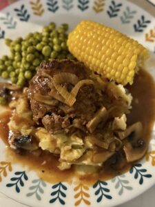 Salisbury steak with potatoes, ear of corn and scoop of peas on a plate.