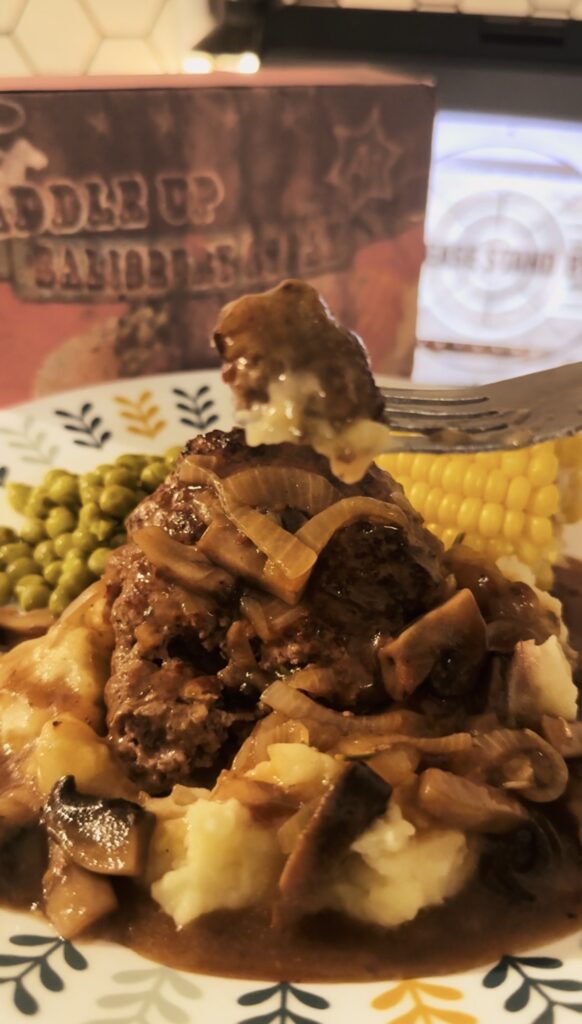 A silver fork holding Salisbury steak smothered in mushroom gravy sauce over mashed potatoes. With corn and peas on the side. A saddle up salisbury tv dinner box in the background.