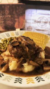 Salisbury steak smothered in mushroom gravy sauce over mashed potatoes. With corn and peas on the side. A saddle up salisbury tv dinner box in the background.