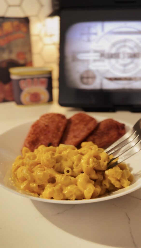 A silver fork scooping up a bit of cheesy mac and cheese noodles. With fried spam in the background.