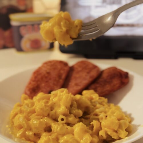 A silver fork scooping up a bit of cheesy mac and cheese noodles. With fried spam in the background.