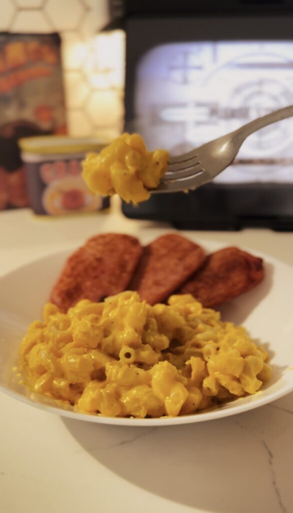 A silver fork scooping up a bit of cheesy mac and cheese noodles. With fried spam in the background.