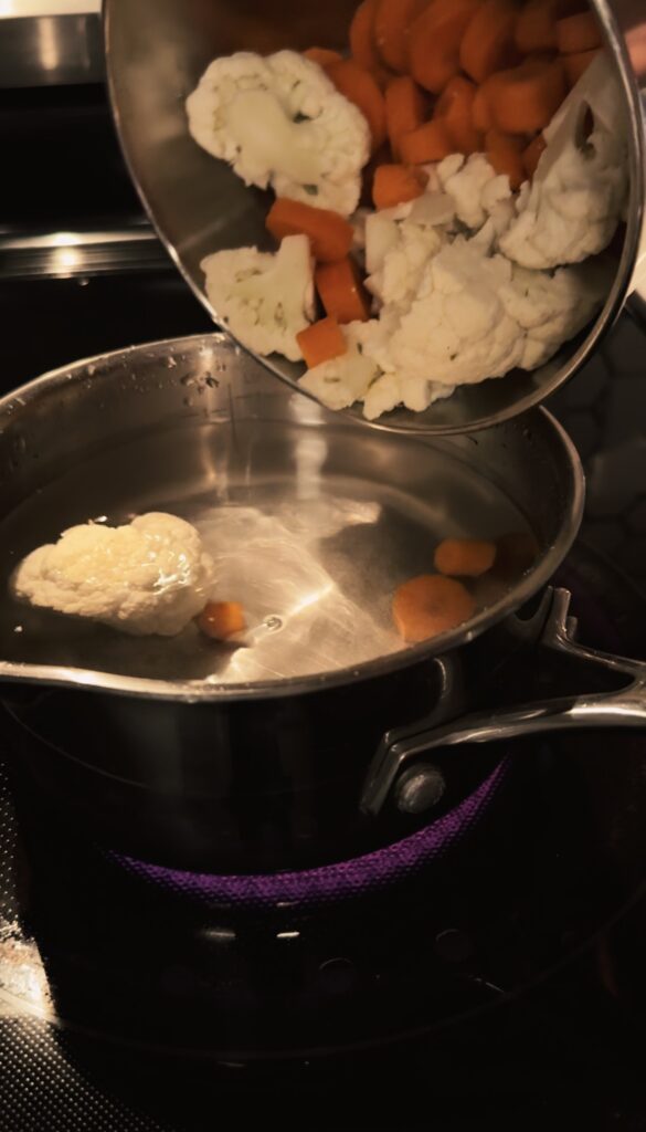 Adding chopped cauliflower and carrots to a pot of boiling water.